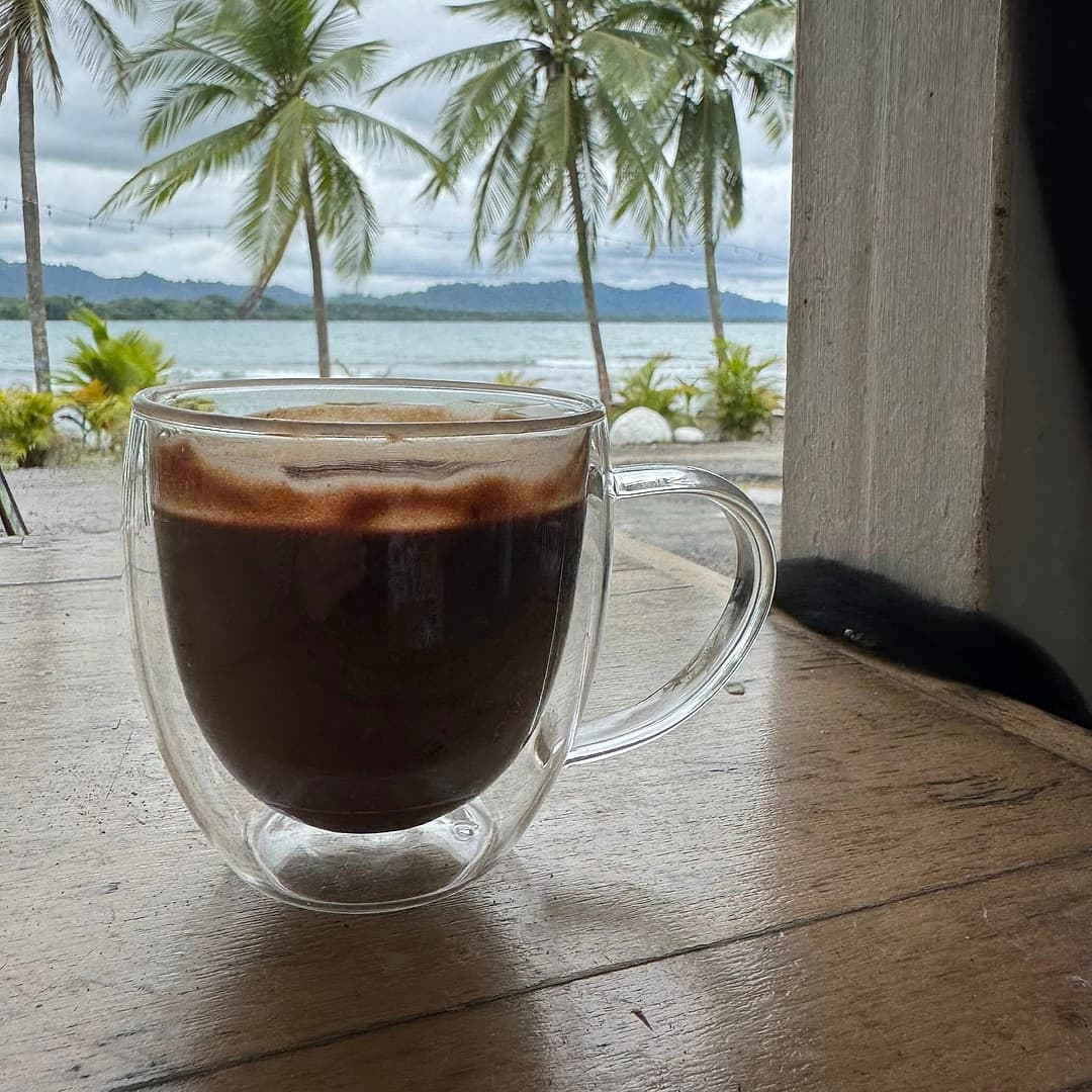A glass mug of hot chocolate on a wooden table with palms and the sea behind, at Cacao Huasi