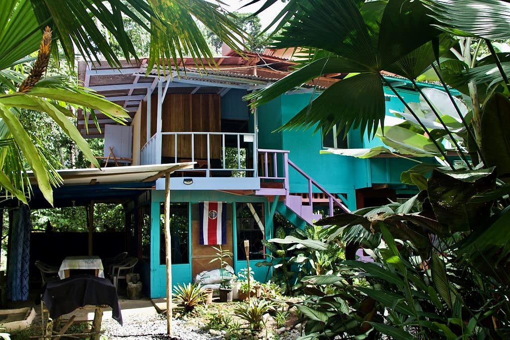 Two-storey turquoise wooden house with an upper balcony, outside staircase and a Costa Rican flag among palms