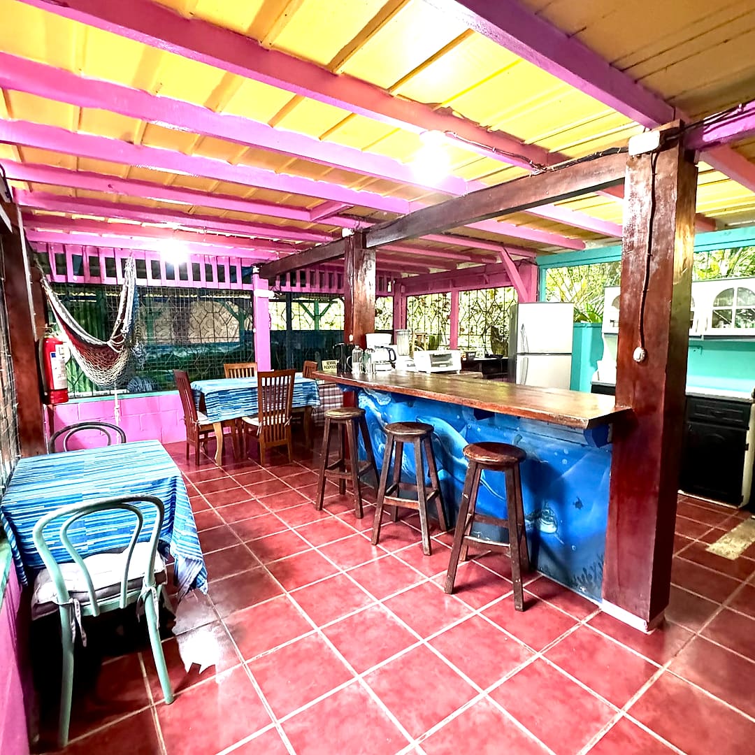 Open-air guest kitchen with a long blue bar counter, wooden stools, pink beams and a yellow ceiling
