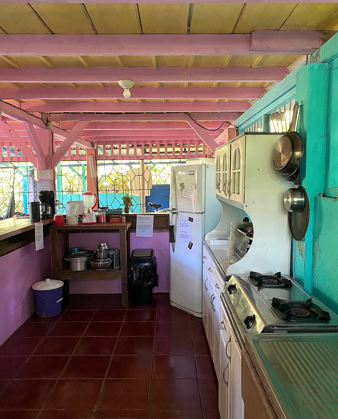 The long open-air guest kitchen under a pink ceiling: stove, hanging pots, fridge and counter