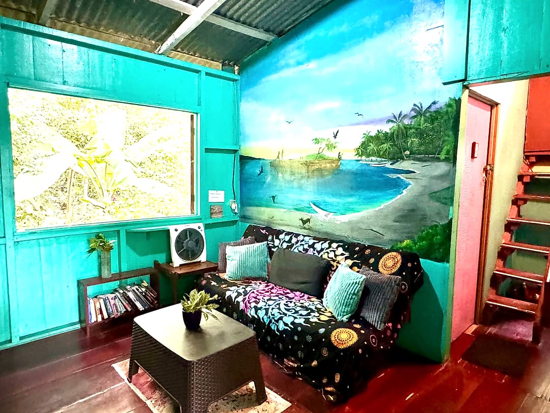 Turquoise lounge with a big painted island-and-bay mural, patterned sofa, wide window and the stairs up