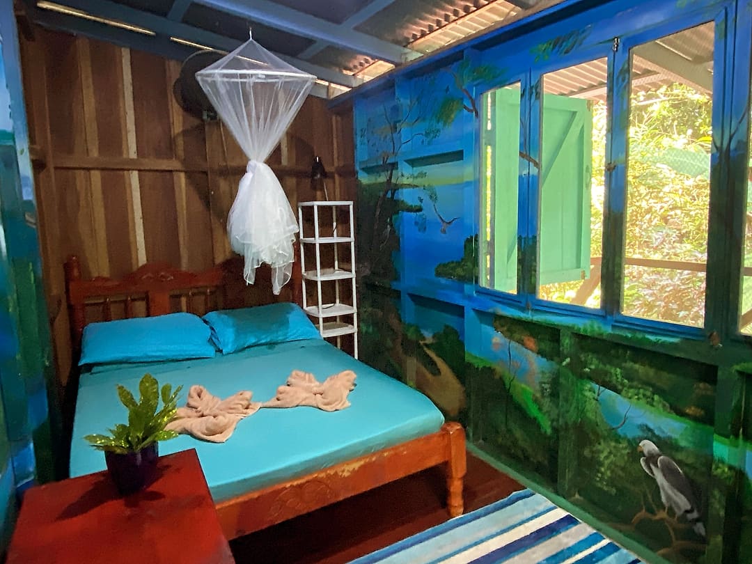 Balcony Room 1: double bed with a turquoise cover under a mosquito net, walls painted with a blue jungle and a heron, green shutters open to the balcony