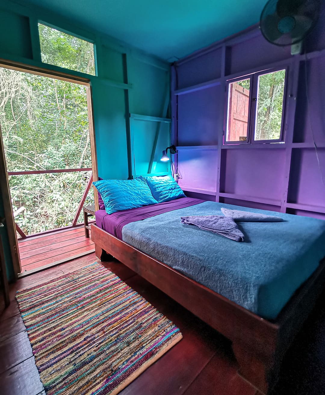 Jungle Balcony Room 4: double bed with a blue cover, violet and turquoise walls, door open to the jungle balcony