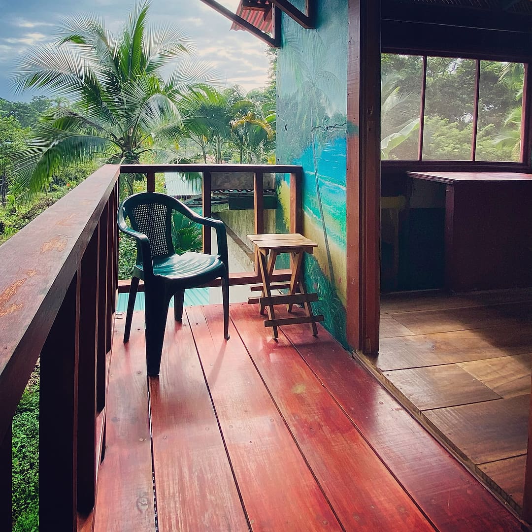 Treetop Balcony Room 5: balcony with a green chair, folding stool and a painted palm-beach wall, looking over palms