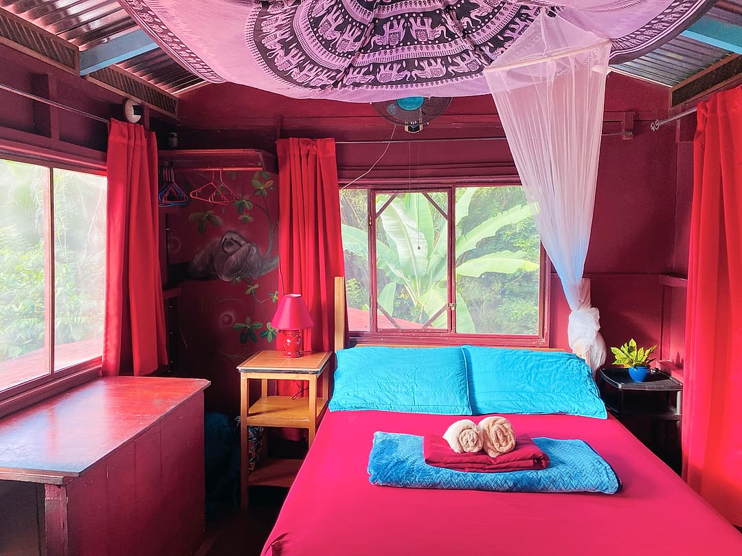 Treetop Balcony Room 5: red walls and curtains, a mandala tapestry on the ceiling, a painted sloth and a bed under a mosquito net